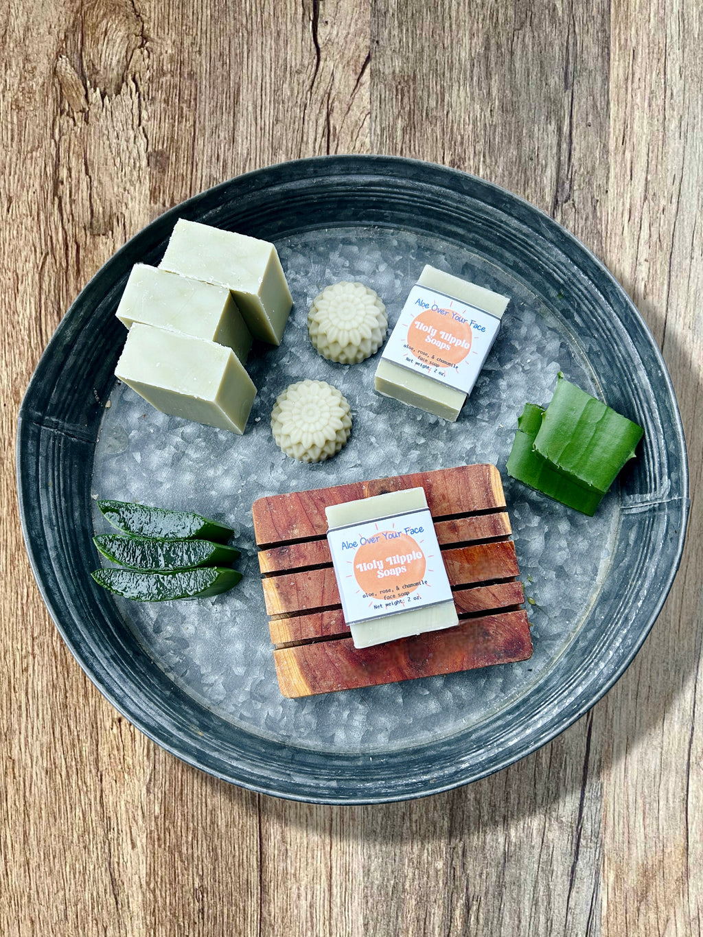 A styled flat-lay of handmade soaps on a dark gray metal tray filled, set on a rustic wooden table. The soaps include pale green rectangular bars, textured pale green round bars, and a wooden soap saver with a pale green soap on top featuring a label the reads Holy Hippie Soaps Aloe Over Your Face. There is sliced aloe in the tray as well.