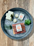 A styled flat-lay of handmade soaps on a dark gray metal tray filled, set on a rustic wooden table. The soaps include pale green rectangular bars, textured pale green round bars, and a wooden soap saver with a pale green soap on top featuring a label the reads Holy Hippie Soaps Aloe Over Your Face. There is sliced aloe in the tray as well.