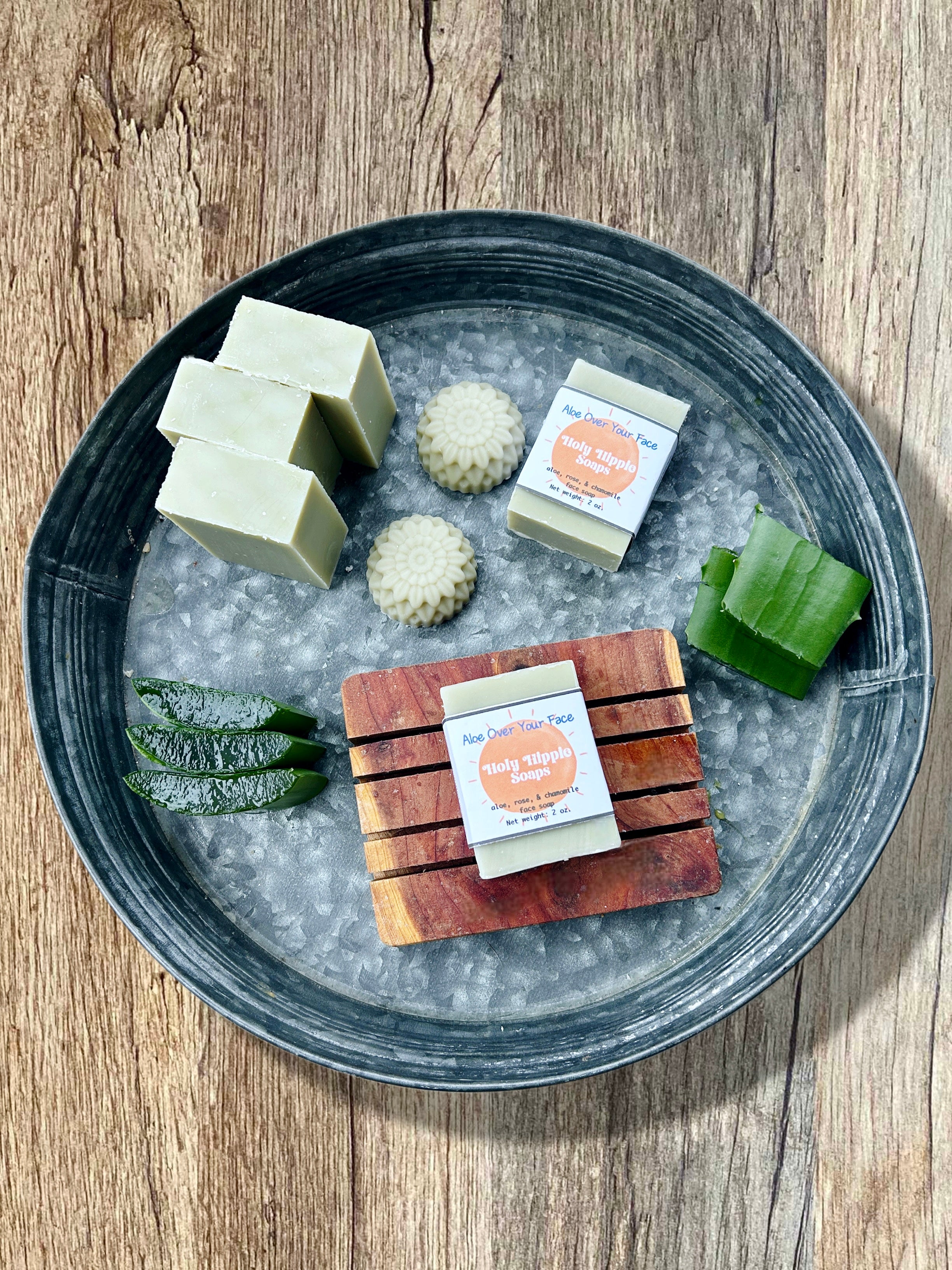 A styled flat-lay of handmade soaps on a dark gray metal tray filled, set on a rustic wooden table. The soaps include pale green rectangular bars, textured pale green round bars, and a wooden soap saver with a pale green soap on top featuring a label the reads Holy Hippie Soaps Aloe Over Your Face. There is sliced aloe in the tray as well.