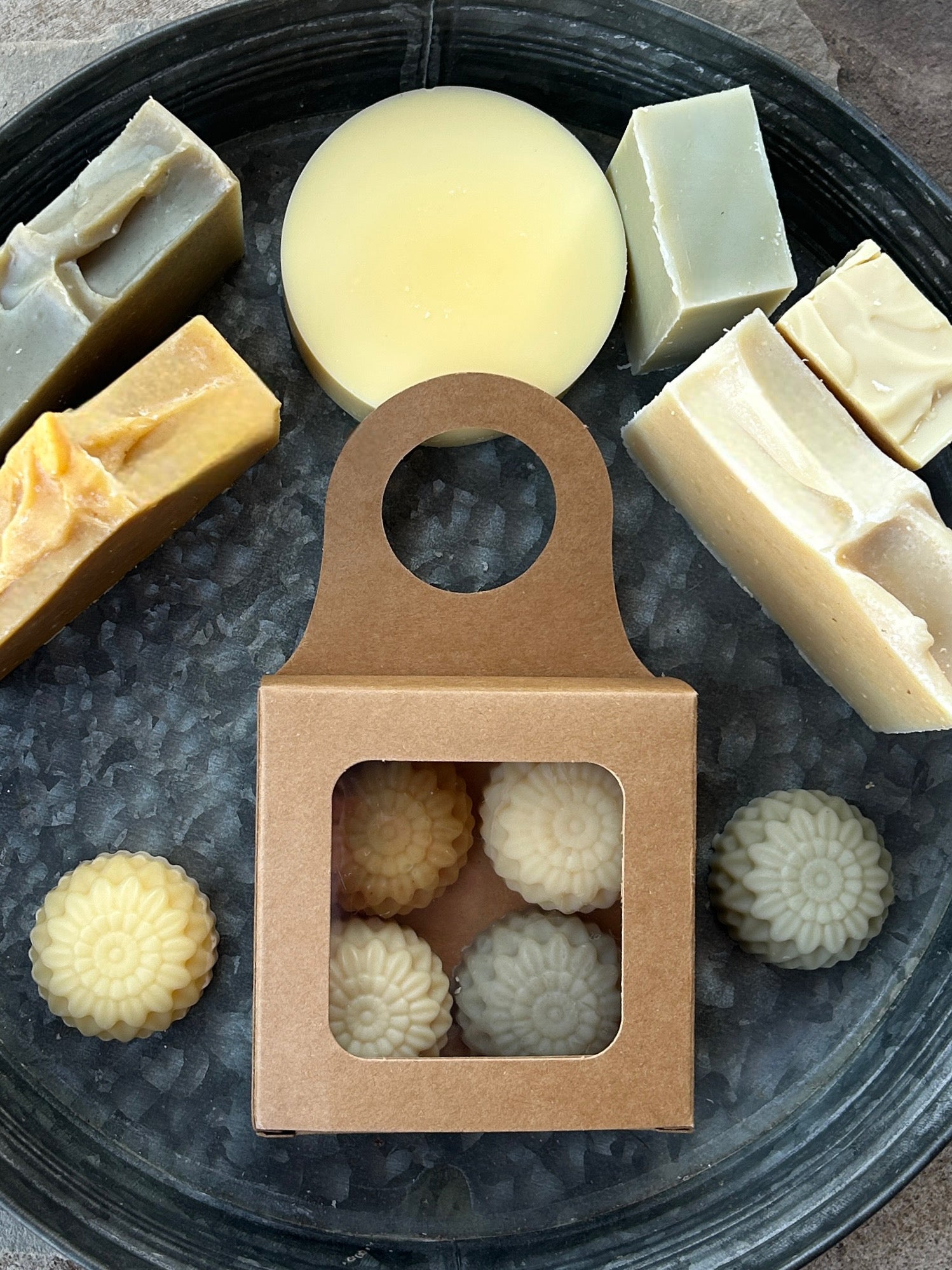 A styled flat-lay of full size handmade soaps as well as round flower sample sized soaps arranged on a dark round tray. In the center is a small kraft paper gift box with a clear window showing four round flower-shaped mini soaps inside.