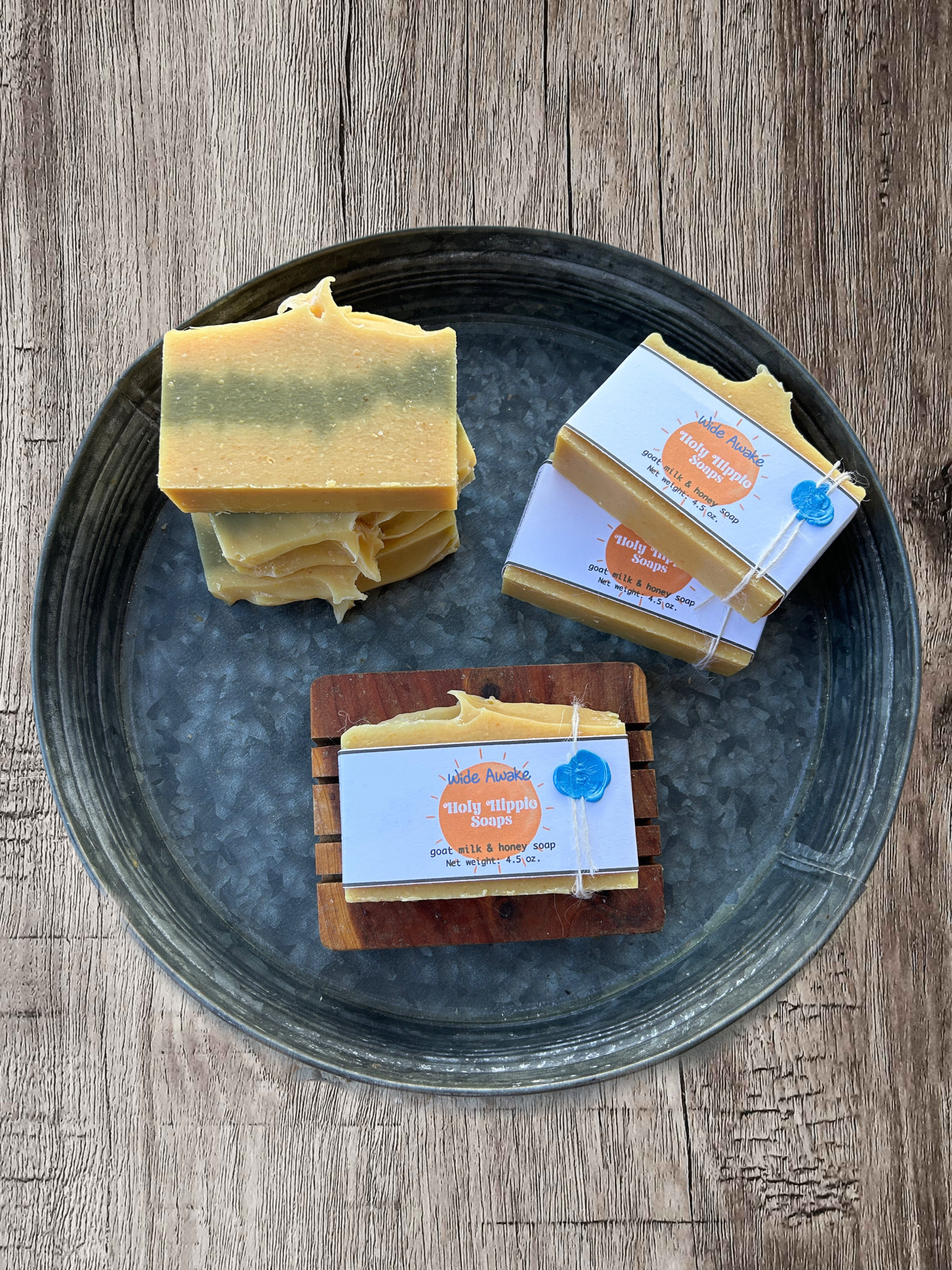 Three stacks of handmade bars of  Wide Awake soap from 'Holy Hippie Soaps' arranged on a round dark gray metal tray on a rustic wooden table. One bar is a rectangular bar with yellow and green marbling, while the other two are wrapped in white white labels with orange and blue branding.