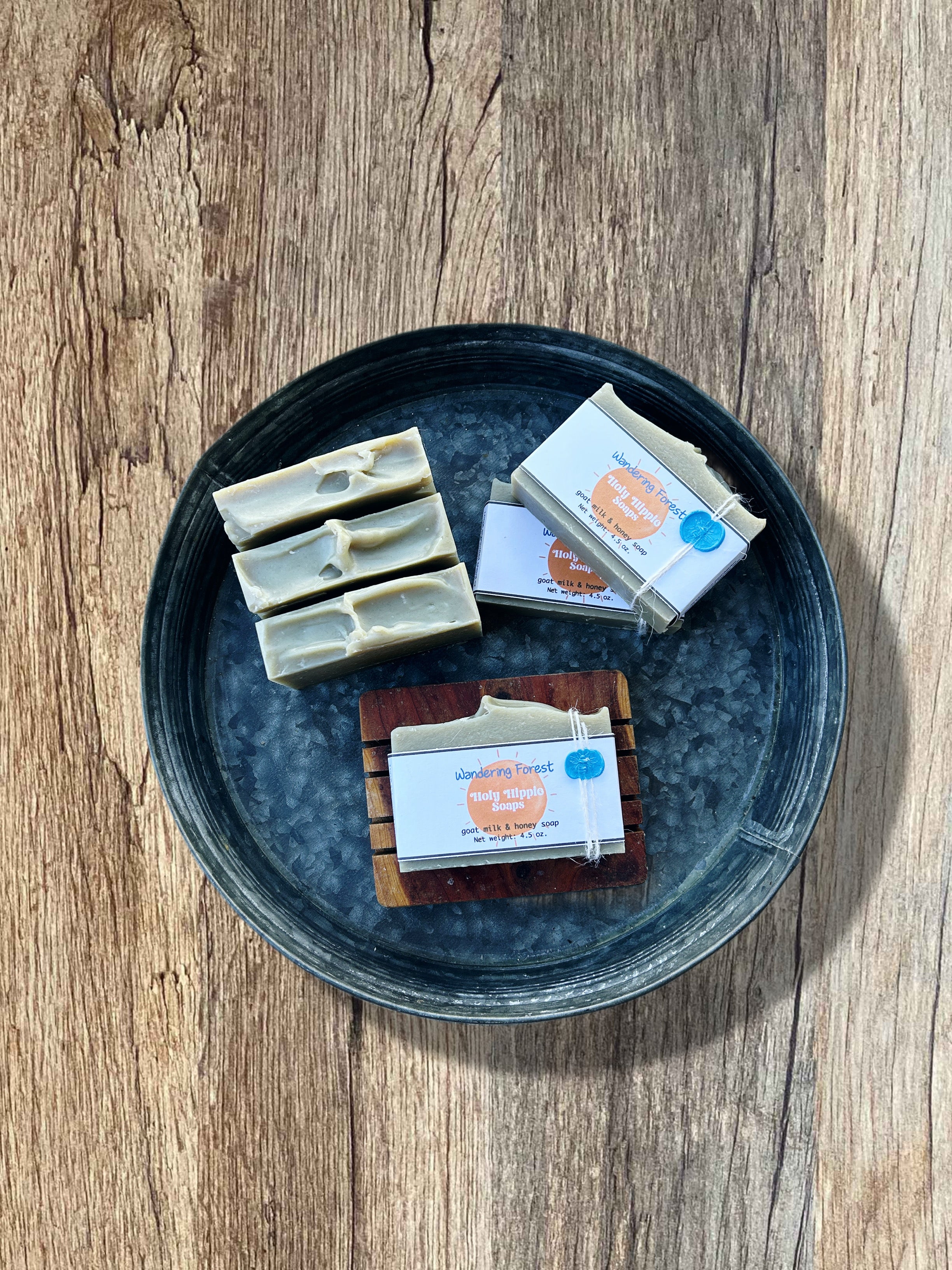 Handmade soap bars displayed on a round metal tray over a rustic wood surface. Three pale green-colored soaps with textured tops are standing up grouped together, and three packaged soap bars with Wandering Forest Holy Hippie Soaps labels are arranged beside them laying flat. Two are laying flat on the tray and the other is on top of a wooden soap saver.