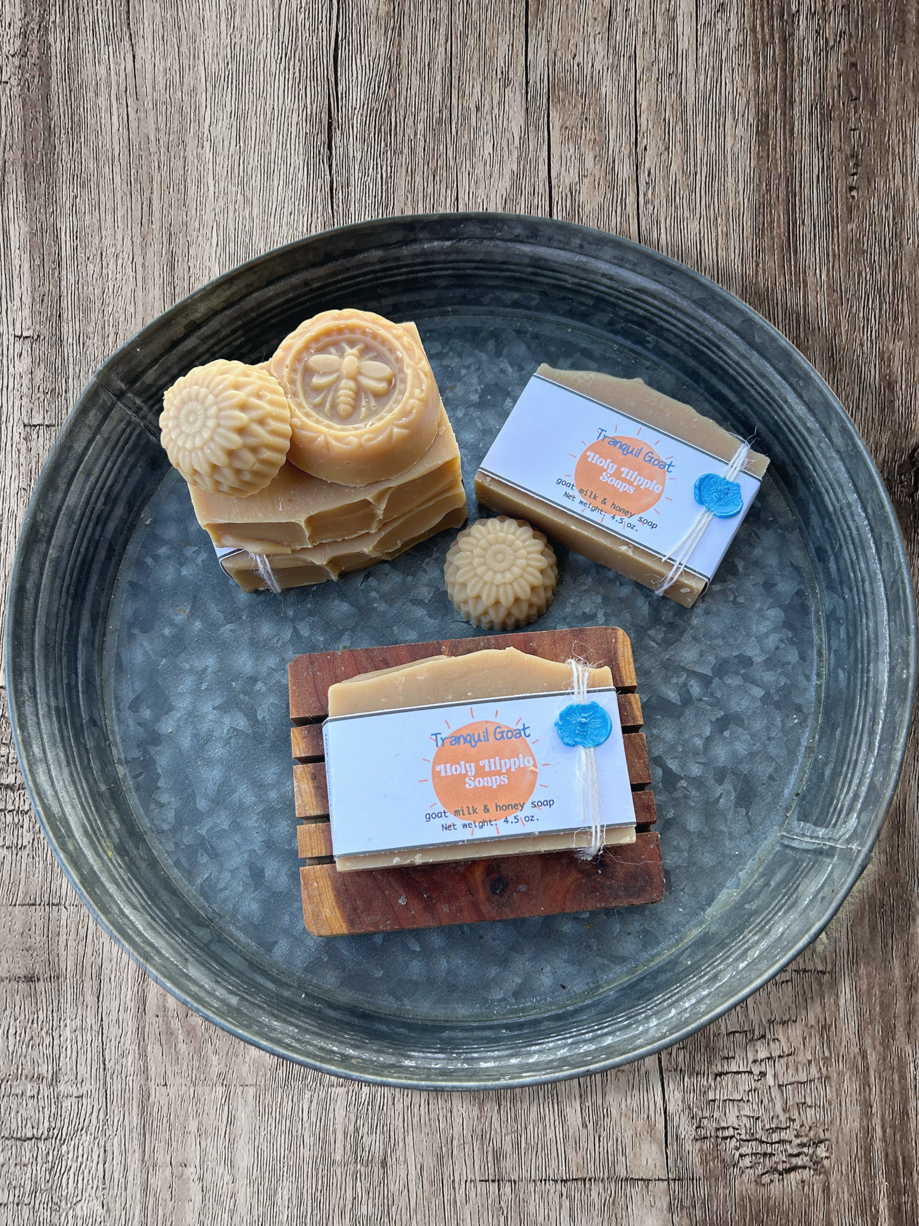 Three bars of handmade beige colored Tranquil Holy Hippie Soap displayed on a rustic dark metal tray over a wooden surface. One bar is stacked with a carved bee design and also a round flower, one is wrapped in a white kraft paper band with a white and orange label, and one sits on a small wooden soap saver with its matching label.