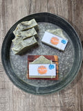Handmade soap bars displayed on a round metal tray over a rustic wood surface. Three green soaps with black speckles are standing up grouped together, and two packaged soap bars with Holy Hippie Soaps labels are arranged beside them laying flat. One is laying flat on the tray and the other is on top of a wooden soap saver.