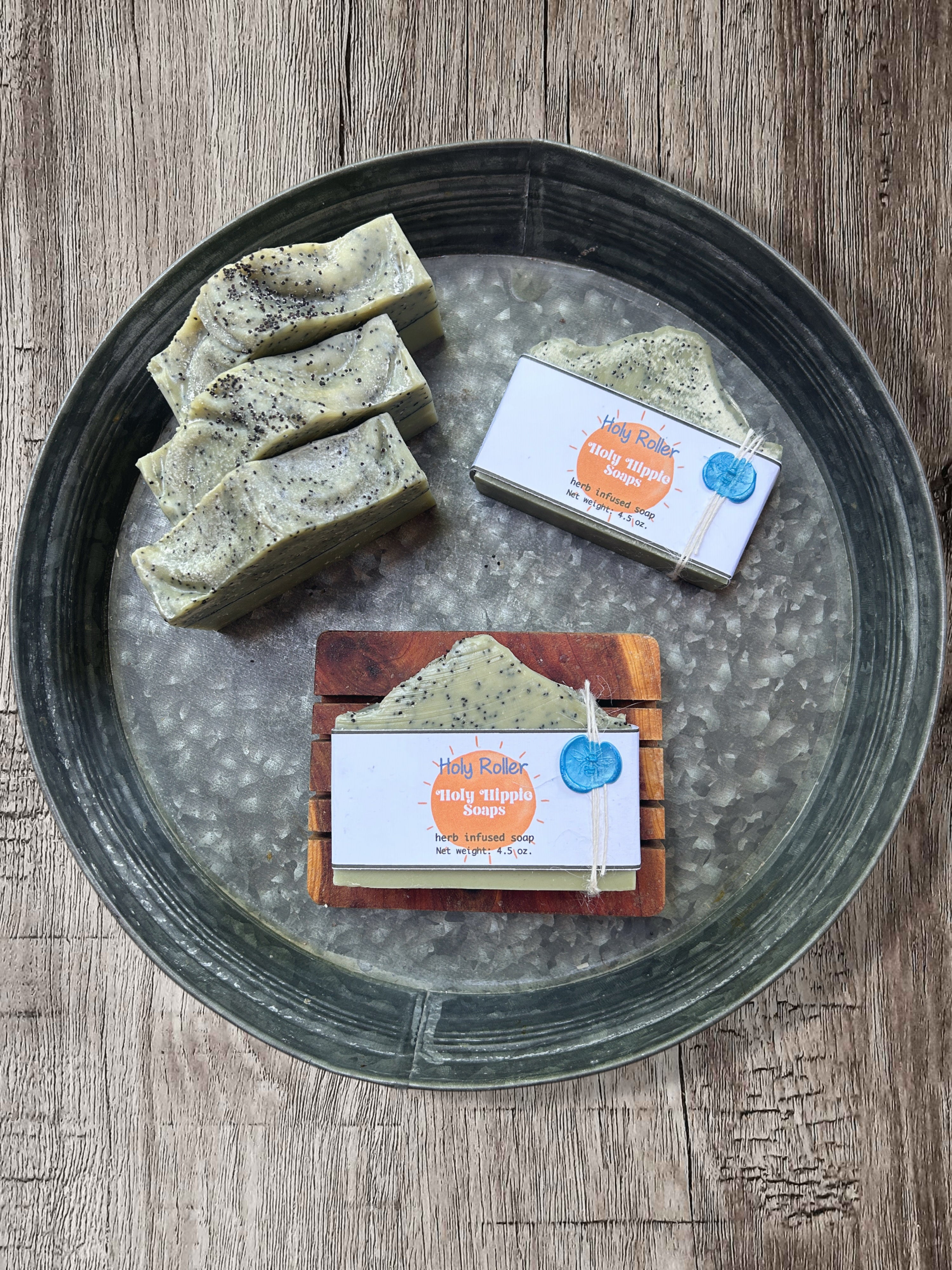 Handmade soap bars displayed on a round metal tray over a rustic wood surface. Three green soaps with black speckles are standing up grouped together, and two packaged soap bars with Holy Hippie Soaps labels are arranged beside them laying flat. One is laying flat on the tray and the other is on top of a wooden soap saver.