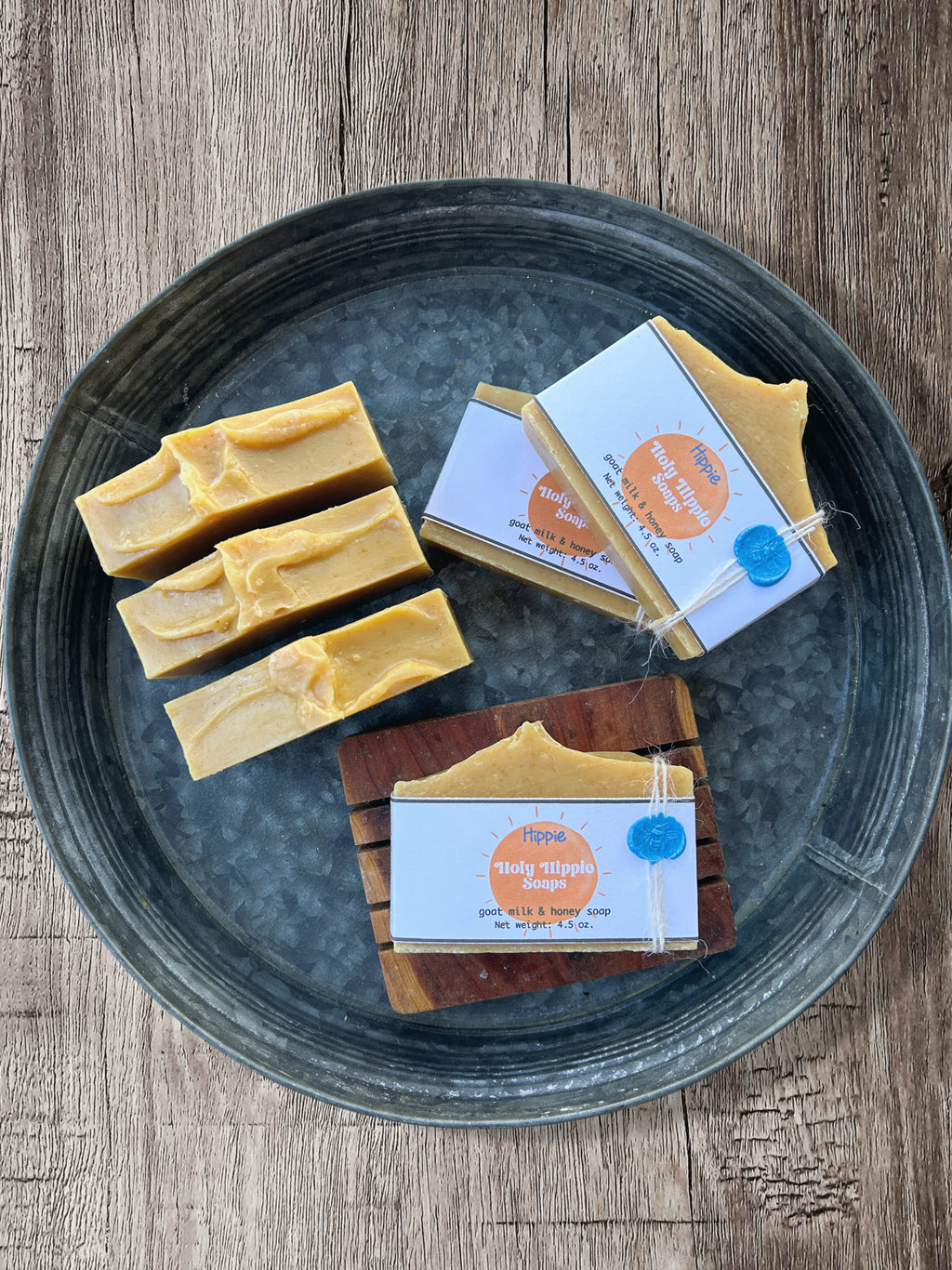 Five bars of handmade soap displayed on a round galvanized metal tray over a rustic wooden surface. Three bars are unwrapped, showing a creamy yellow texture, while two are wrapped in white labels that read "Holy Hippie Soaps Hippie". One soap is displayed on a wooden soap saver with the same label.
