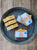 Five bars of handmade soap displayed on a round galvanized metal tray over a rustic wooden surface. Three bars are unwrapped, showing a creamy yellow texture, while two are wrapped in white labels that read "Holy Hippie Soaps Hippie". One soap is displayed on a wooden soap saver with the same label.