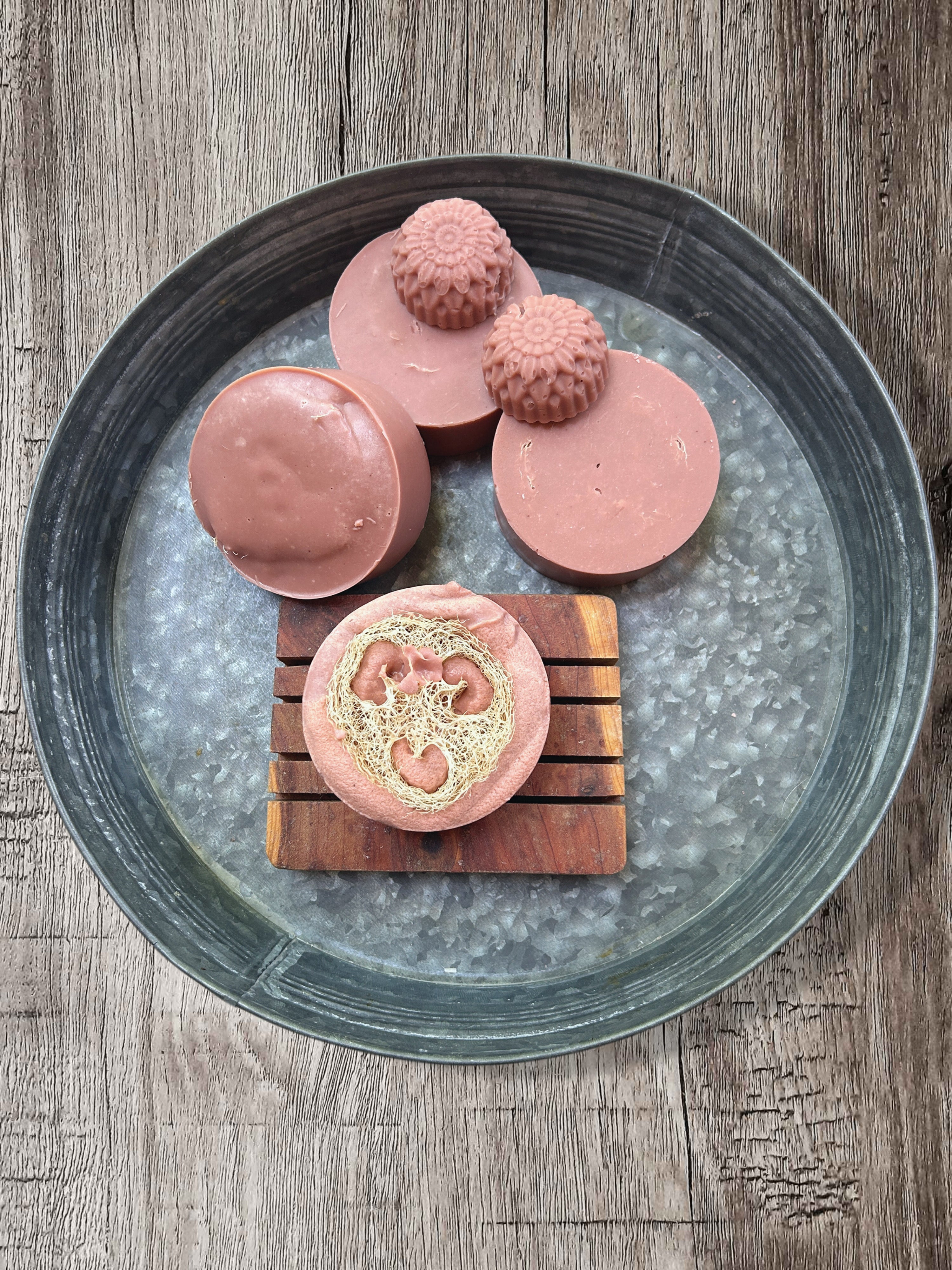 Four handmade pink soap bars arranged on a galvanized metal tray over a rustic wooden surface. One round bar shows the luffa sponge poking out of the soap, while three others are solid pink.