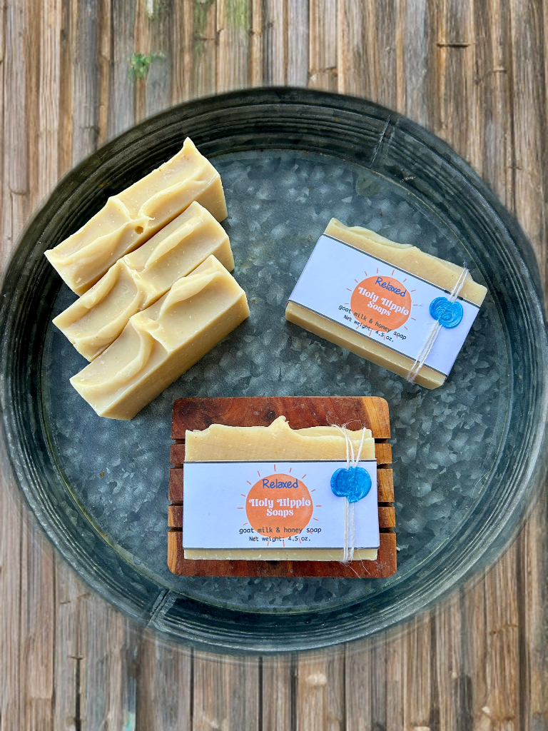 Three bars of handcrafted soap in pale yellowish cream, arranged on a round rustic metal tray on a wooden surface. Two bars are wrapped in a white label with orange and blue details, one bar is placed on a small wooden soap saver and the other is laying flat on the tray displaying Holy Hippie Soaps Relaxed.