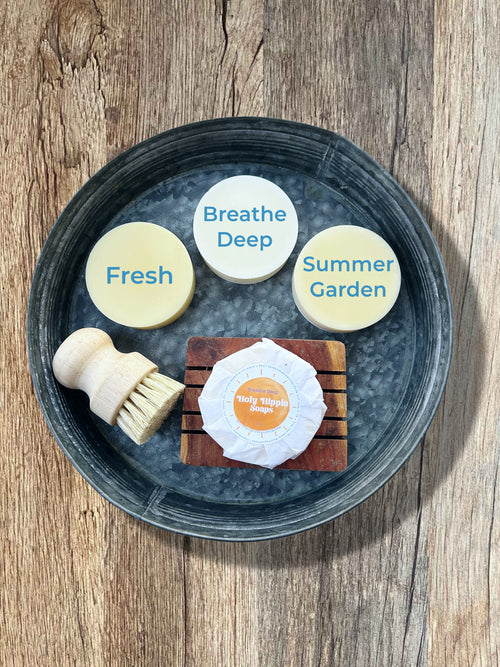 A top-down view of dish soaps arranged on a round dark tray over a wooden surface. The picture includes three round soap bars labeled Fresh, Breathe Deep, and Summer Garden, a wrapped bar labeled Holy Hippie Soaps, a small wooden brush, and a small wooden soap saver.