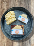 Handmade soap bars displayed on a round metal tray over a rustic wood surface. Three cream-colored soaps with textured tops are standing up grouped together, and two packaged soap bars with Divine Holy Hippie Soaps labels are arranged beside them laying flat. One is laying flat on the tray and the other is on top of a wooden soap saver.