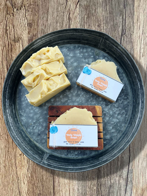 Handmade soap bars displayed on a round metal tray over a rustic wood surface. Three cream-colored soaps with textured tops are standing up grouped together, and two packaged soap bars with Divine Holy Hippie Soaps labels are arranged beside them laying flat. One is laying flat on the tray and the other is on top of a wooden soap saver.
