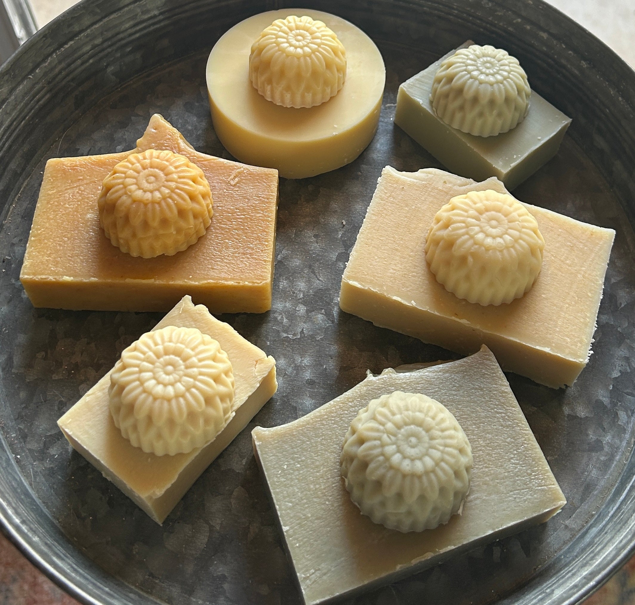 Six bars of handmade soap arranged on a dark metal tray, each topped with a small, circular, flower-patterned soap piece. The soaps come in pale yellow, light beige, pale green, and off-white shades.