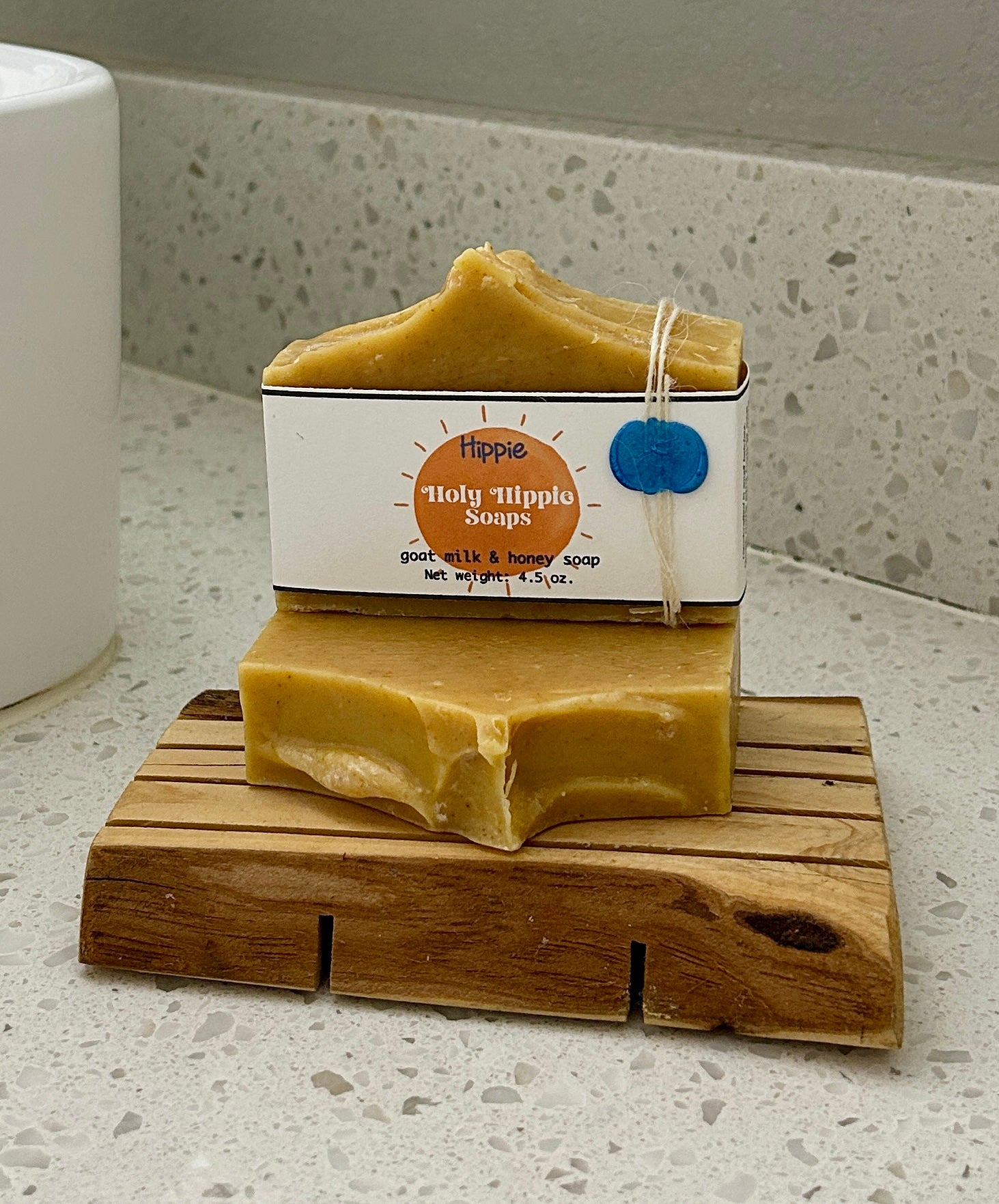 Two bars of handmade Holy Hippie Soaps on a wooden soap saver on a white speckled countertop, with one bar wrapped in a white paper sleeve showing the brand label Hippie soap.