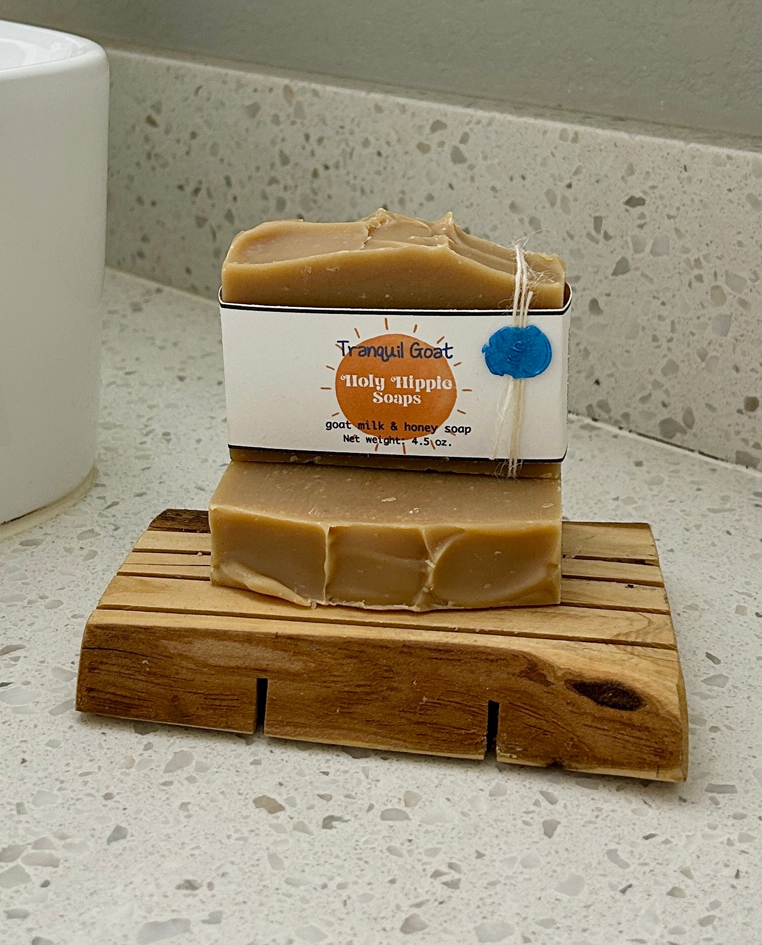 Two bars of beige handmade soap labeled 'Tranquil Holy Hippie Soap'  sits on a wooden soap saver on a white speckled countertop, with a white sink partially visible on the left.
