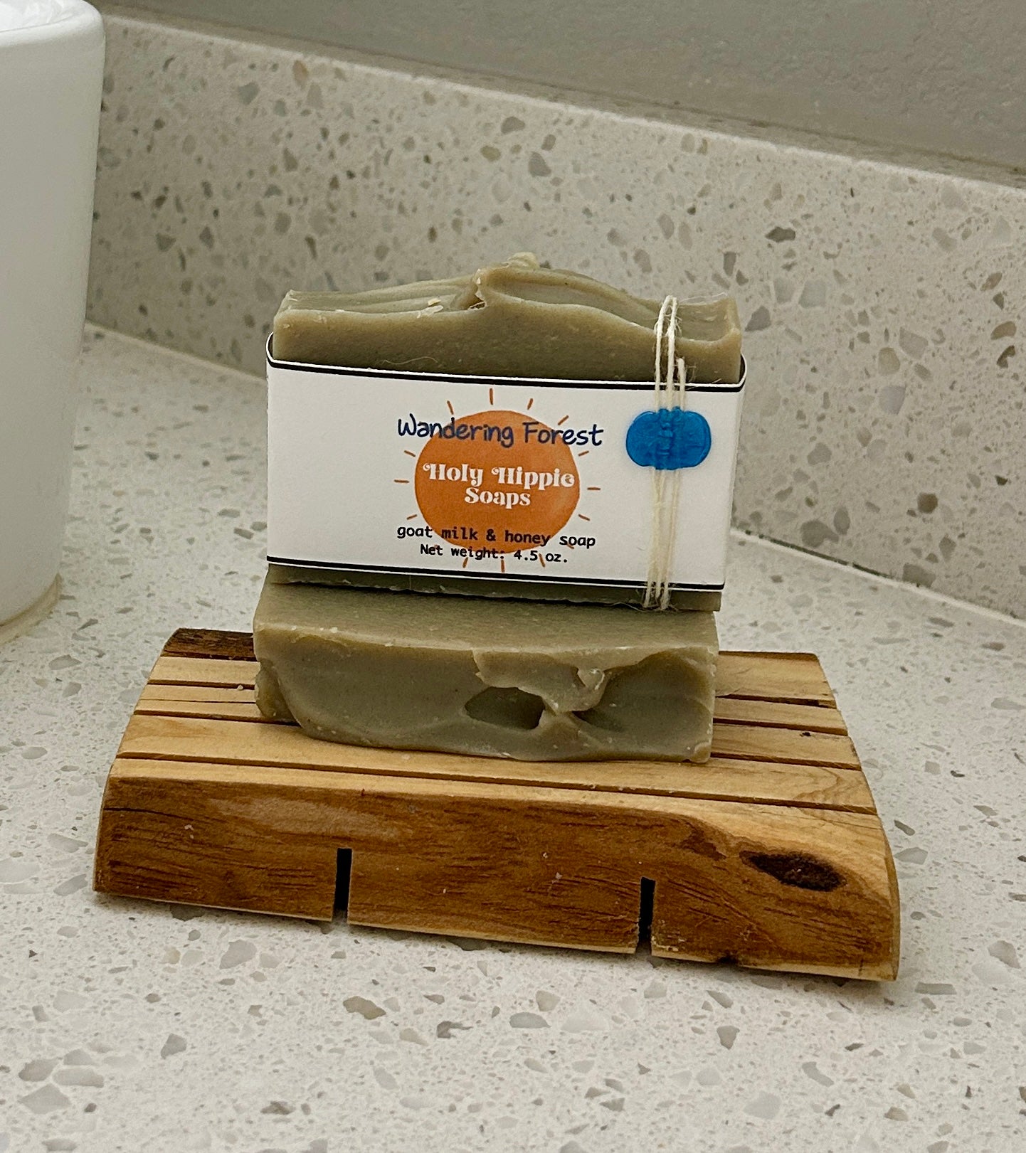 Two bars of pale green handmade soap from Wandering Forest Holy Hippie Soaps resting on a wooden soap saver on a white speckled countertop next to a white sink.