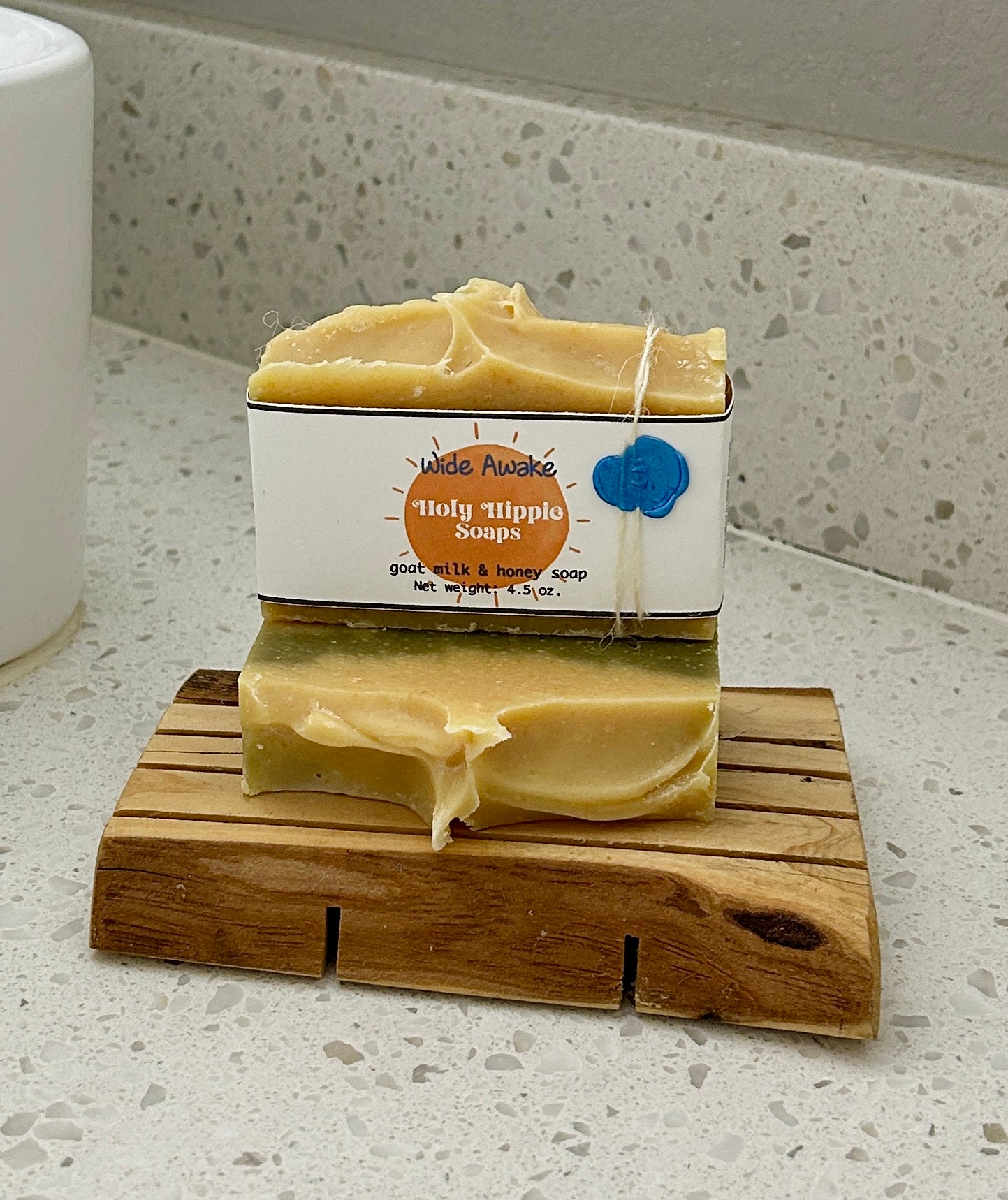 Two bars of handmade Wide Awake soap from Holy Hippie Soaps displayed on a wooden soap saver on a white speckled countertop next to a white sink.