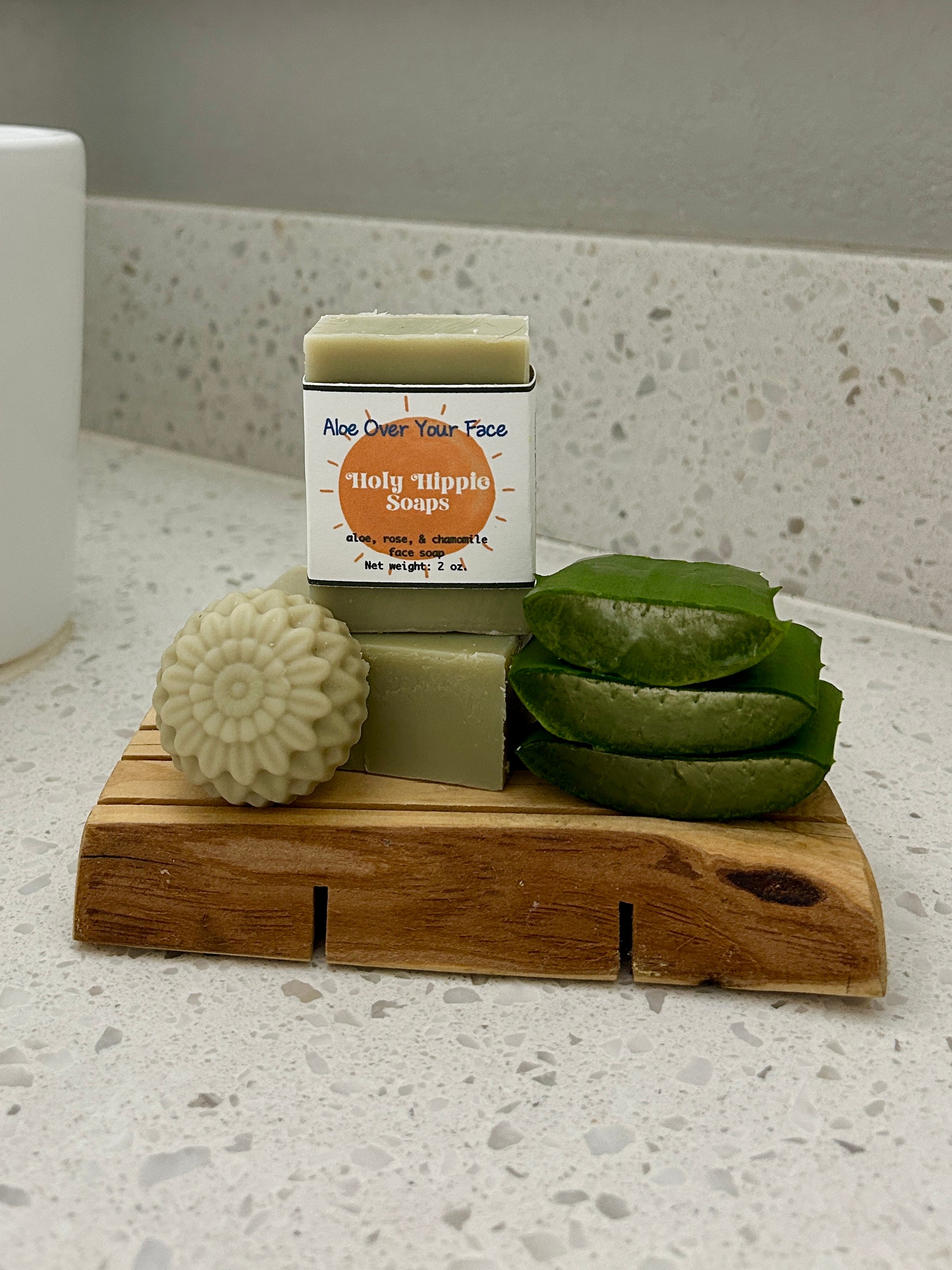 A pale green bar of soap labeled 'Holy Hippie Soap' stands on a wooden soap saver with a round flower soap and green aloe vera slices on a speckled countertop.