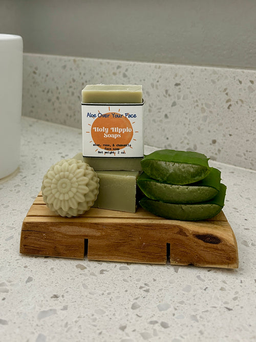 A pale green bar of soap labeled 'Holy Hippie Soap' stands on a wooden soap saver with a round flower soap and green aloe vera slices on a speckled countertop.