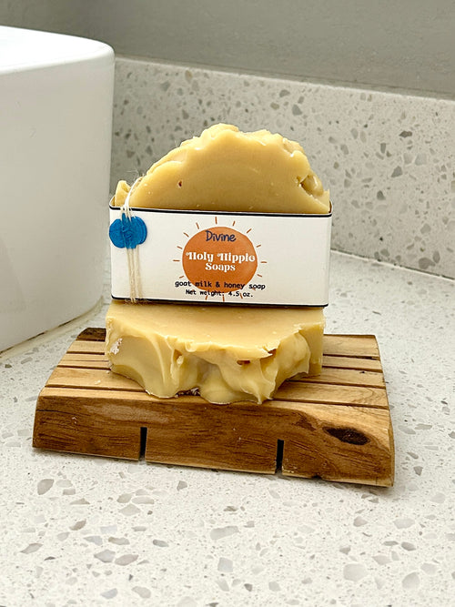 A bar of handmade soap labeled 'Divine Holy Hippie Soaps' resting on a small wooden soap saver on a white speckled countertop next to a white sink.