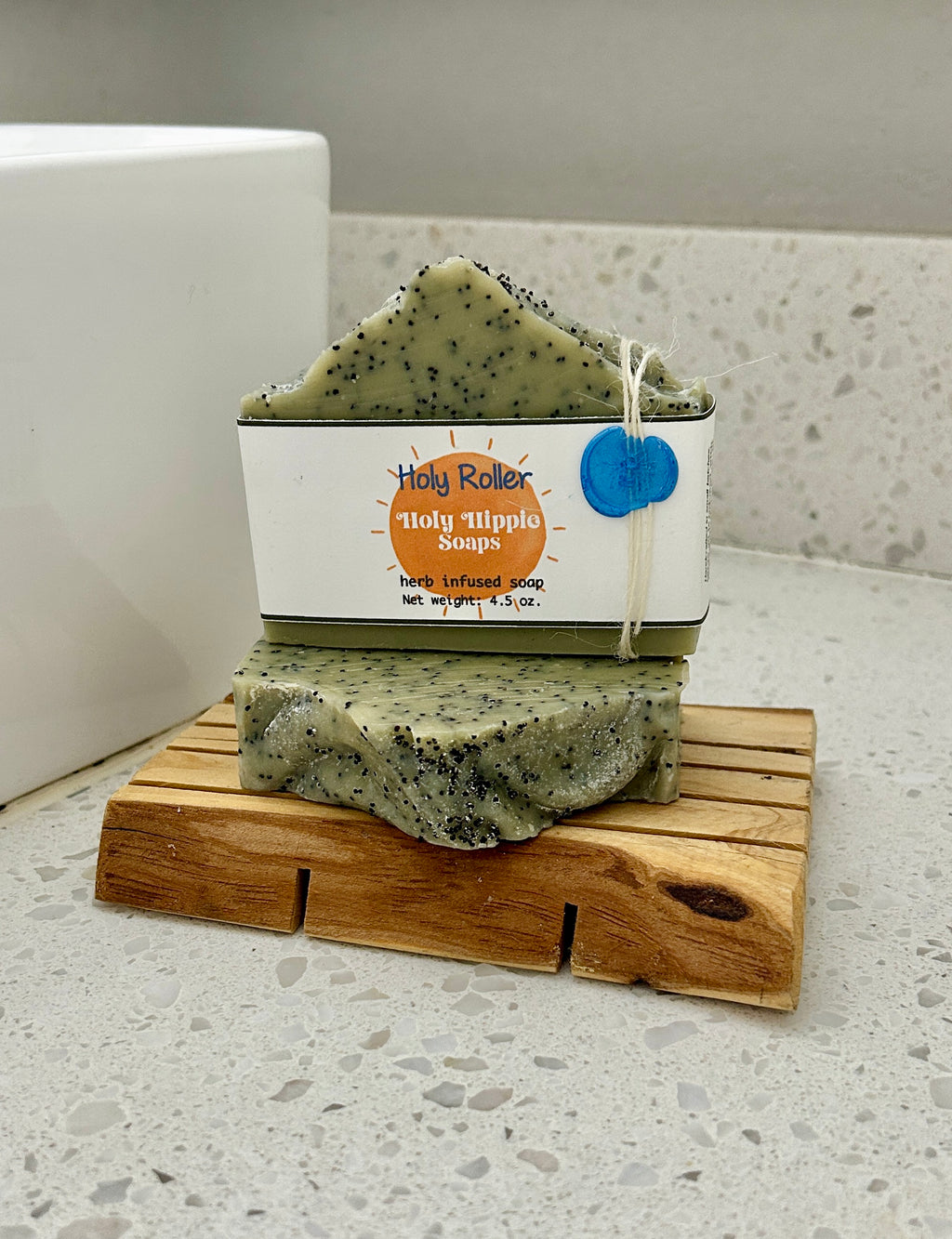 Two bars of Holy Roller Holy Hippie Soap are resting on a small wooden soap saver on a white speckled countertop next to a white sink. The bars of soap are pale green with black speckles.