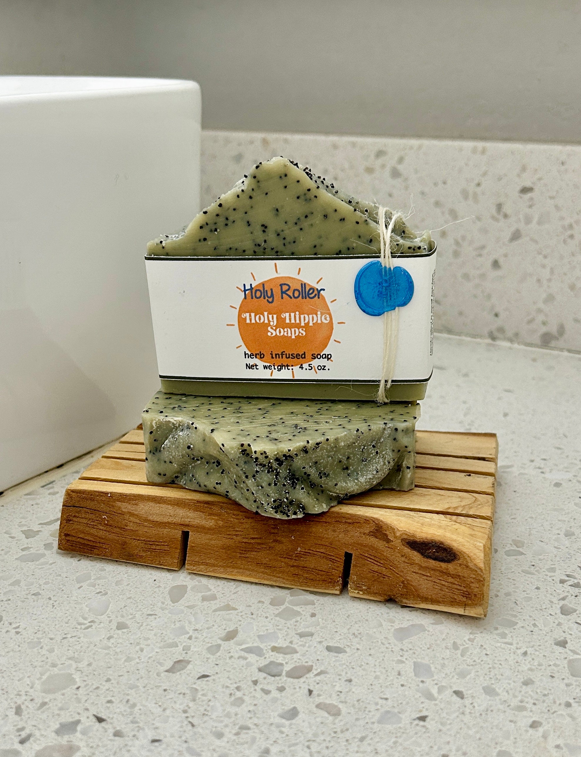 Two bars of Holy Roller Holy Hippie Soap are resting on a small wooden soap saver on a white speckled countertop next to a white sink. The bars of soap are pale green with black speckles.