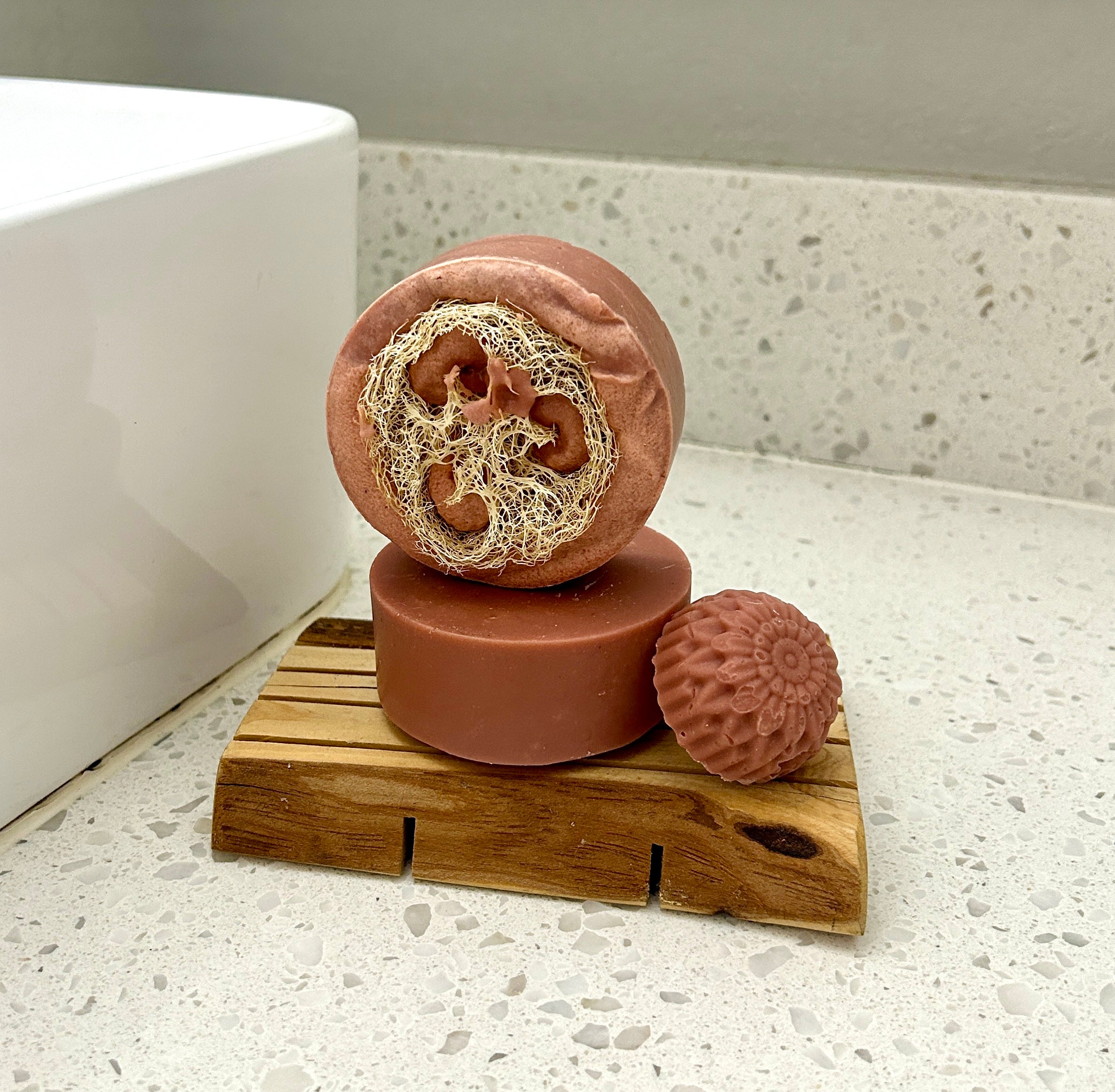 Three rose-colored handmade soap bars displayed on top a small wooden soap saver on a speckled white countertop next to a white sink.