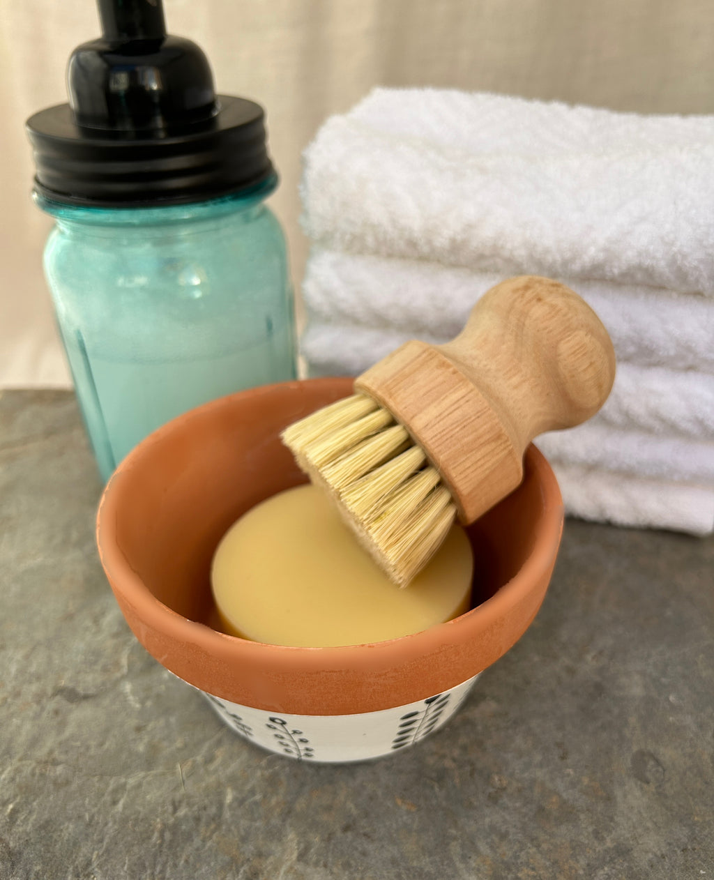 A natural wood-handled bristle brush resting in a small terracotta bowl filled with pale yellow soap, with a folded white towel and a blue glass bottle in the background.