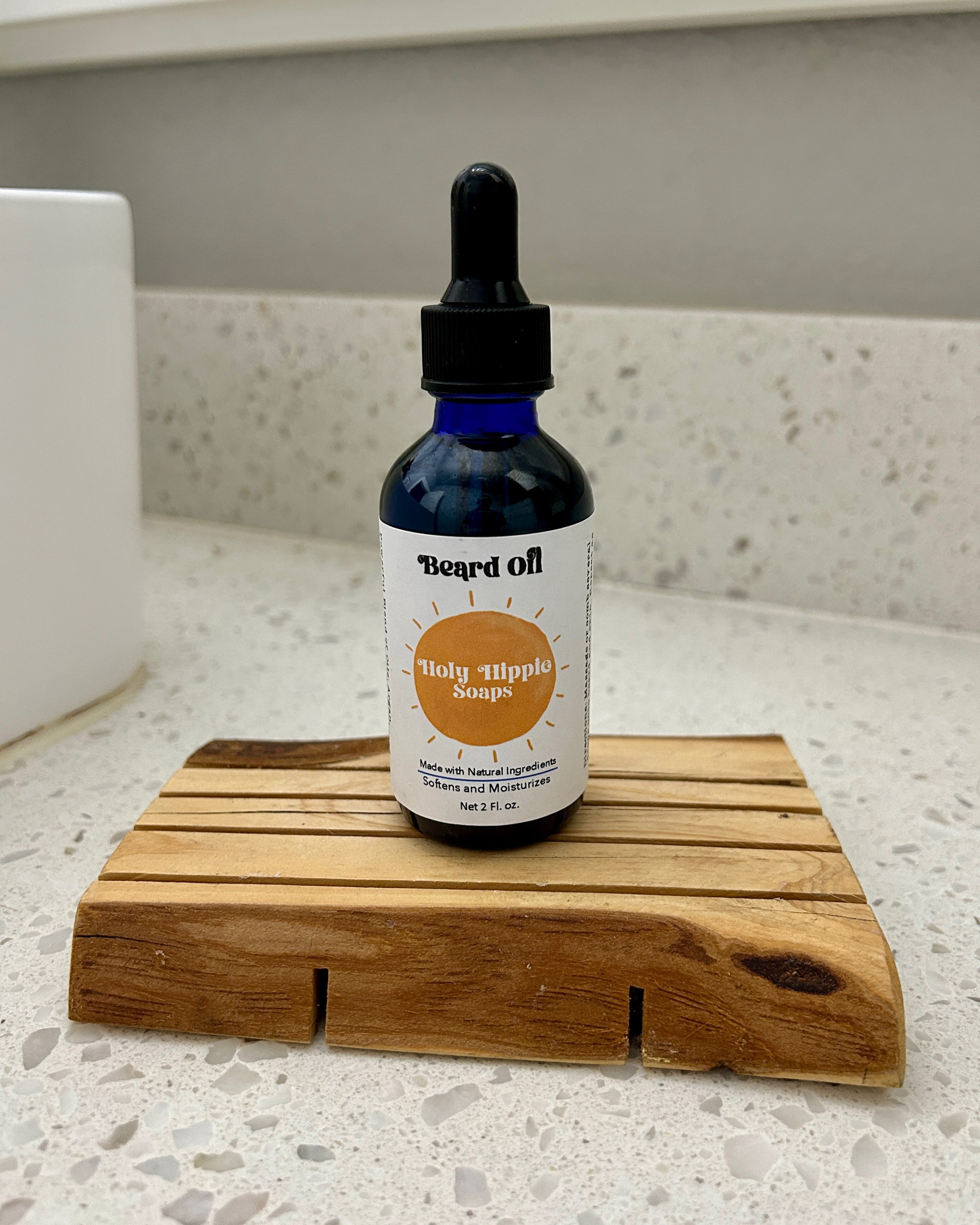 small dropper bottle labeled beard oil on a rustic wooden soap saver sitting on top of a white speckled countertop next to a sink