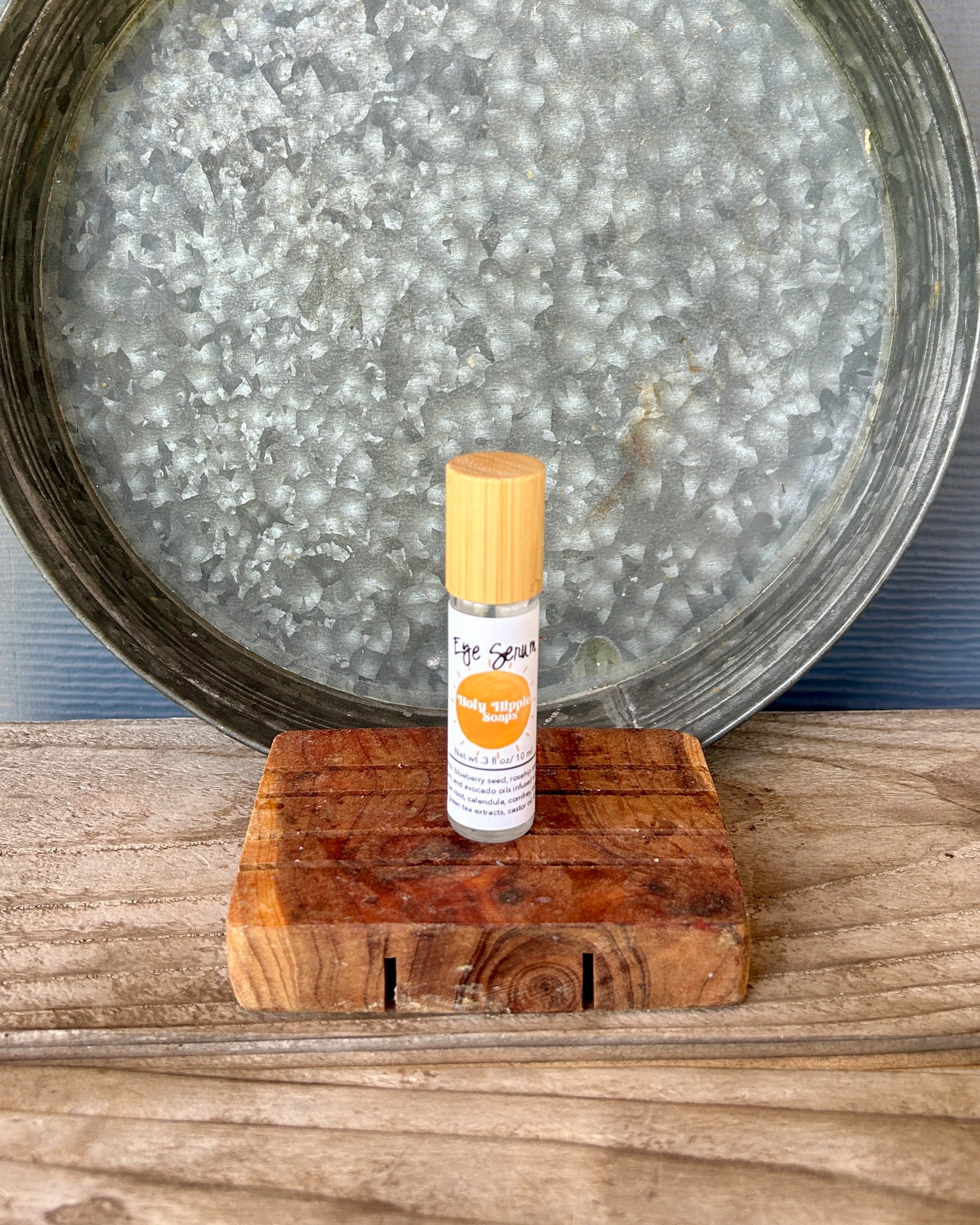 A small roll-on bottle labeled 'Eye Serum' with a light wood cap, displayed on a rustic wooden block in front of a large galvanized metal tray on a wooden surface.
