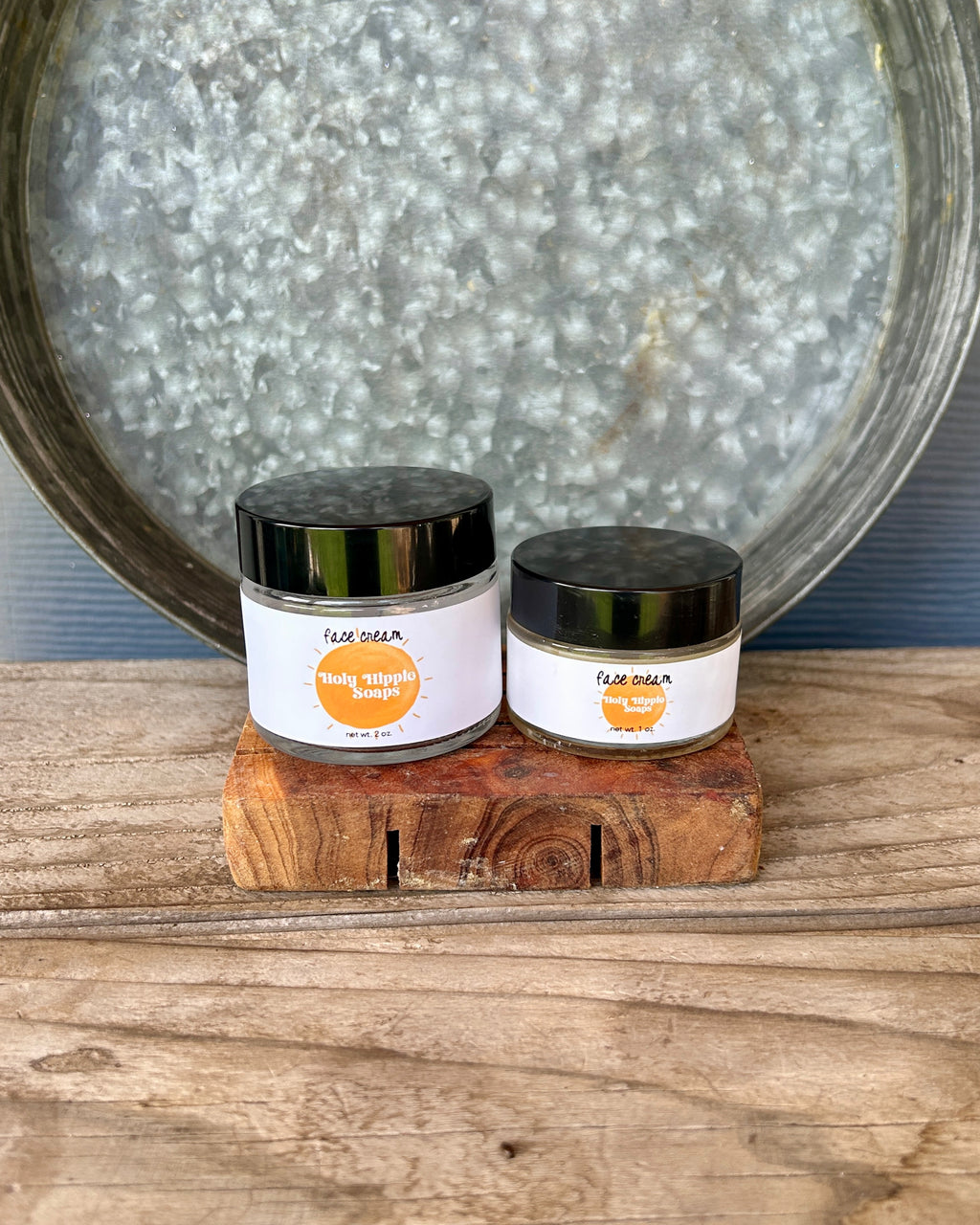 Two small jars of face cream from the brand Holy Hippie Soaps, labeled 'face cream', displayed on a rustic wooden block in front of a galvanized metal tray on a wooden surface.
