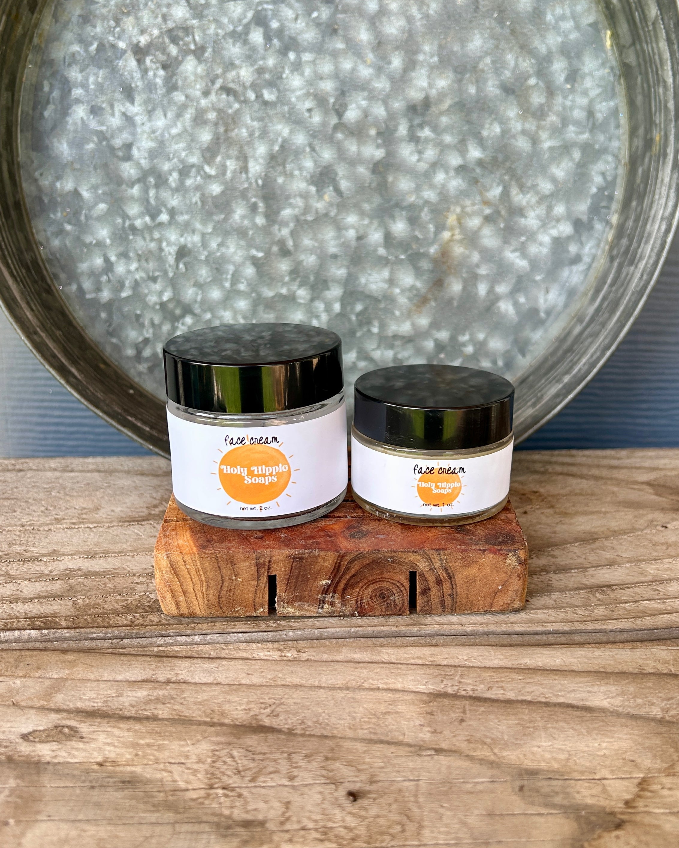 Two small jars of face cream from the brand Holy Hippie Soaps, labeled 'face cream', displayed on a rustic wooden block in front of a galvanized metal tray on a wooden surface.