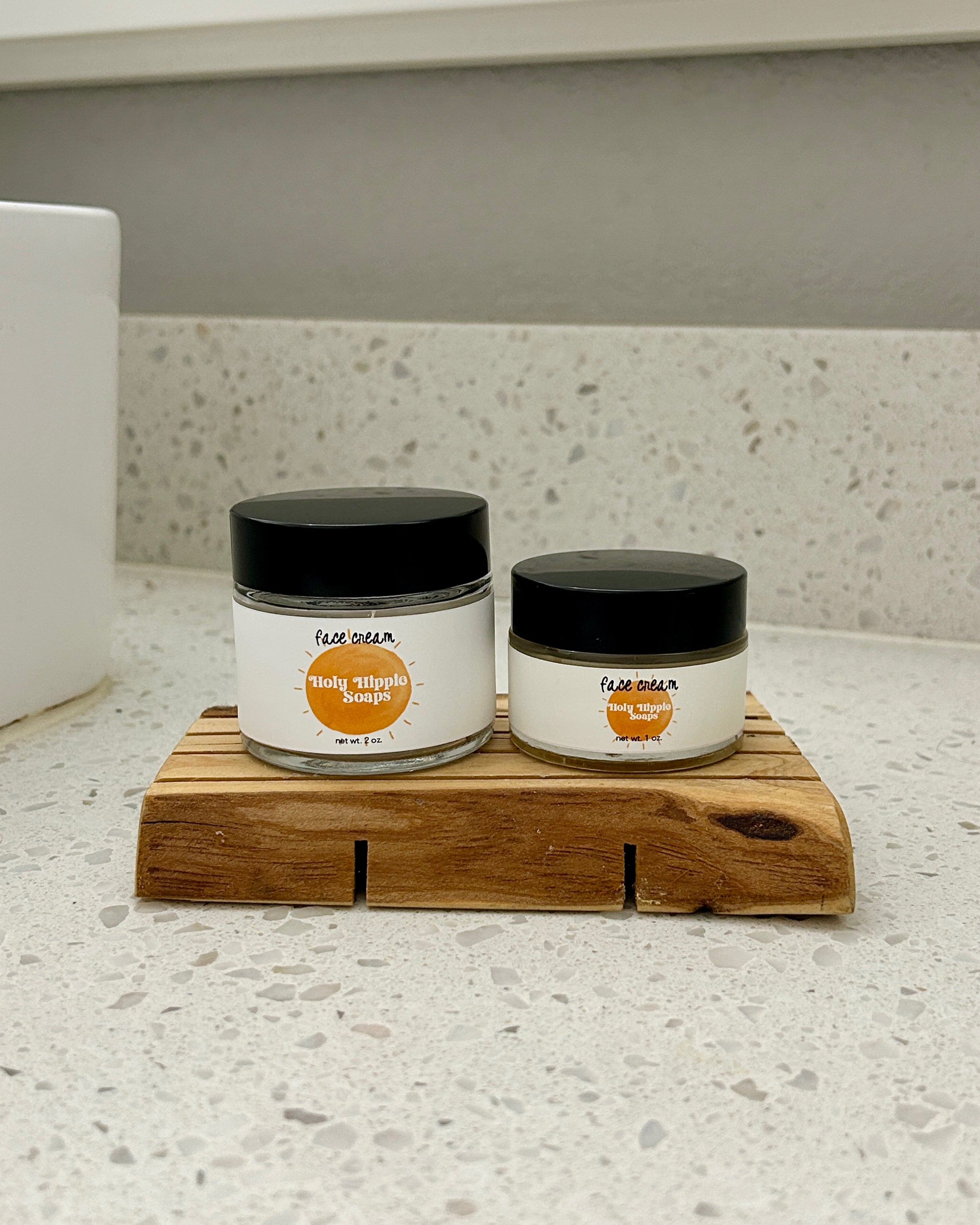 Two small jars of Holy Hippie Soaps face cream on a rustic wooden soap saver on a white speckled countertop.
