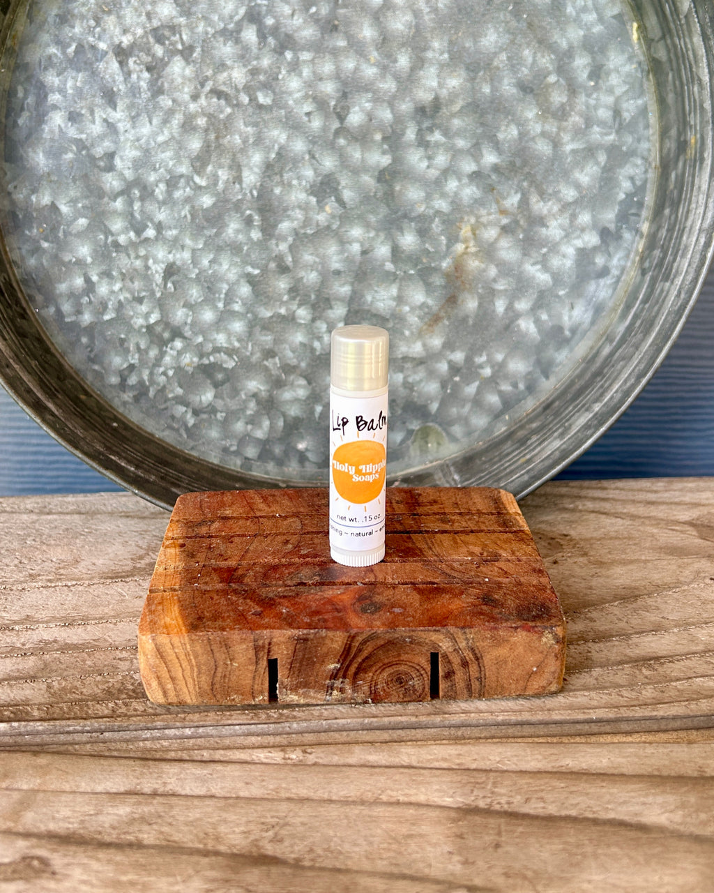 A small tube of lip balm labeled 'Lip Balm' sits on a rustic wooden soap saver, with a large galvanized metal tray in the background.