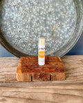 A small tube of lip balm labeled 'Lip Balm' sits on a rustic wooden soap saver, with a large galvanized metal tray in the background.