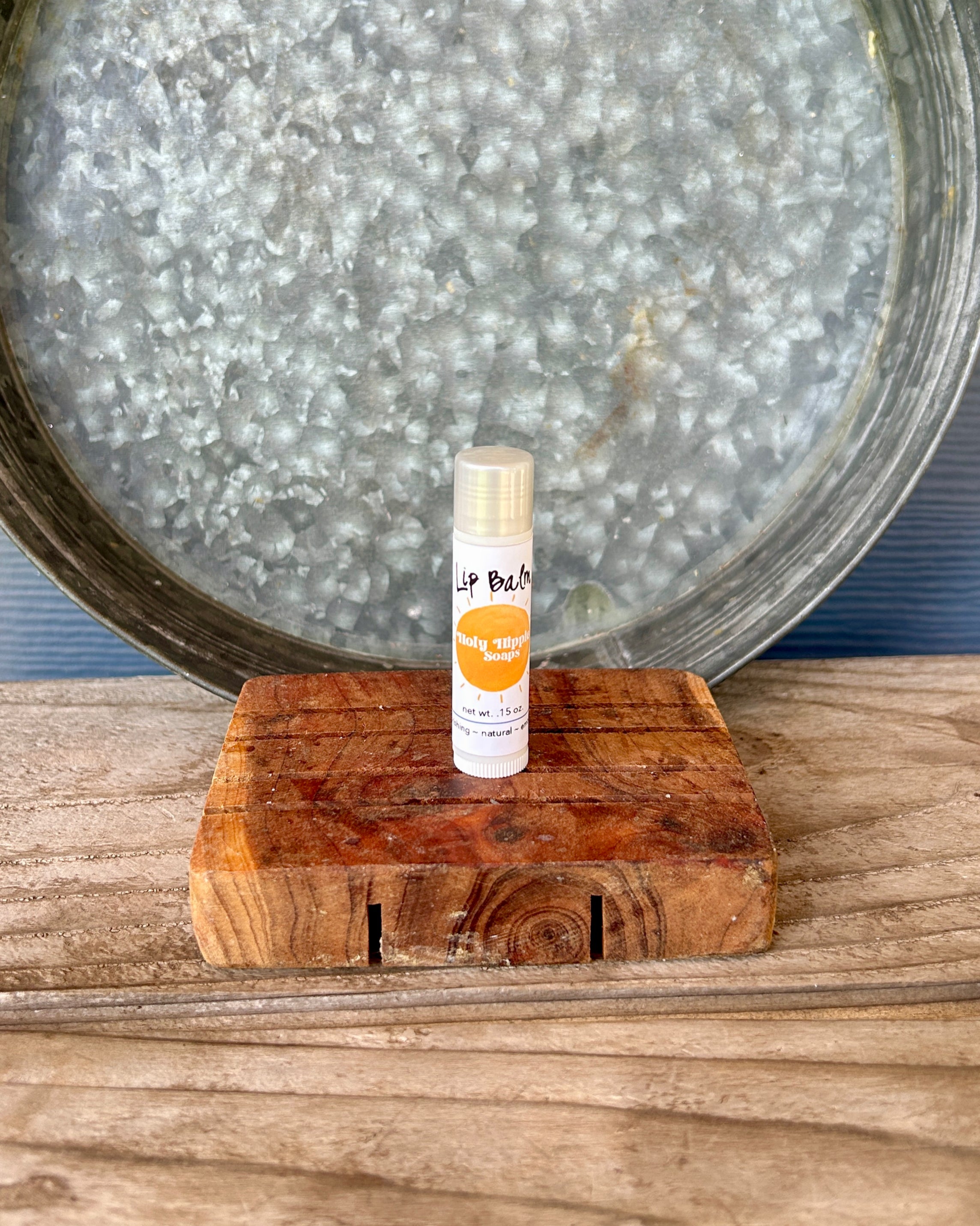 A small tube of lip balm labeled 'Lip Balm' sits on a rustic wooden soap saver, with a large galvanized metal tray in the background.