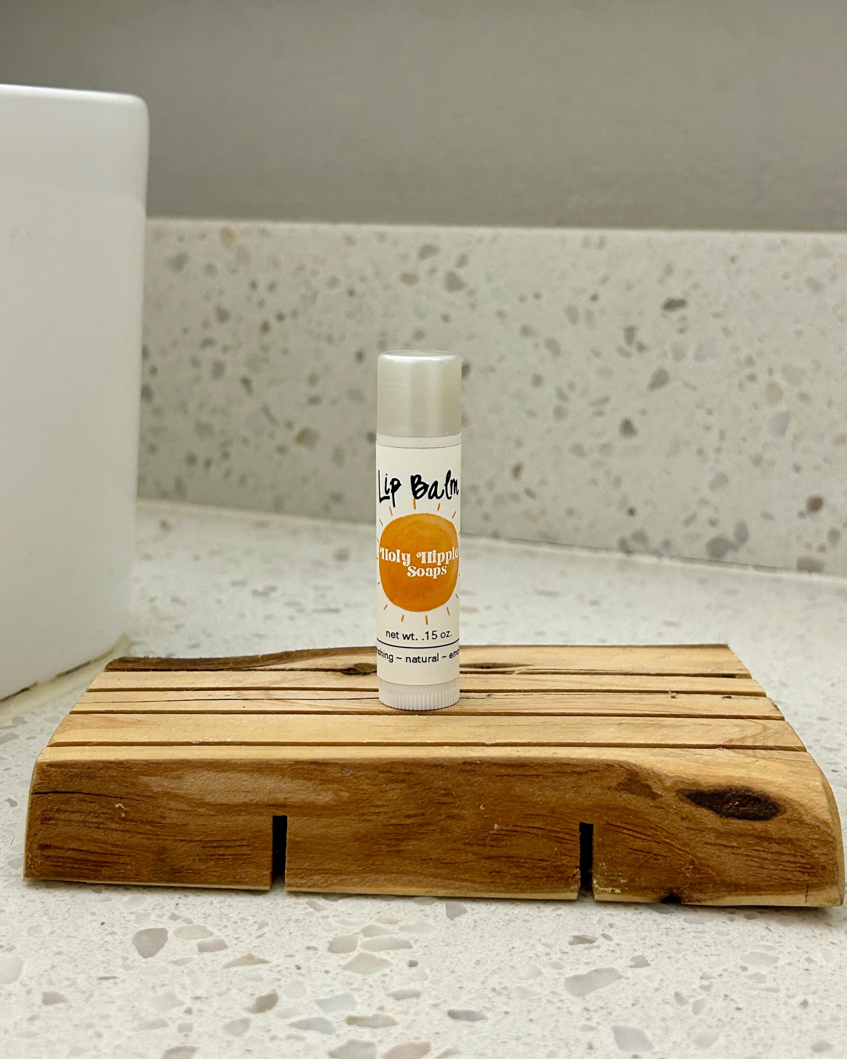 A small tube of lip balm standing upright on a wooden tray on a speckled countertop, with a white sink edge visible in the background.
