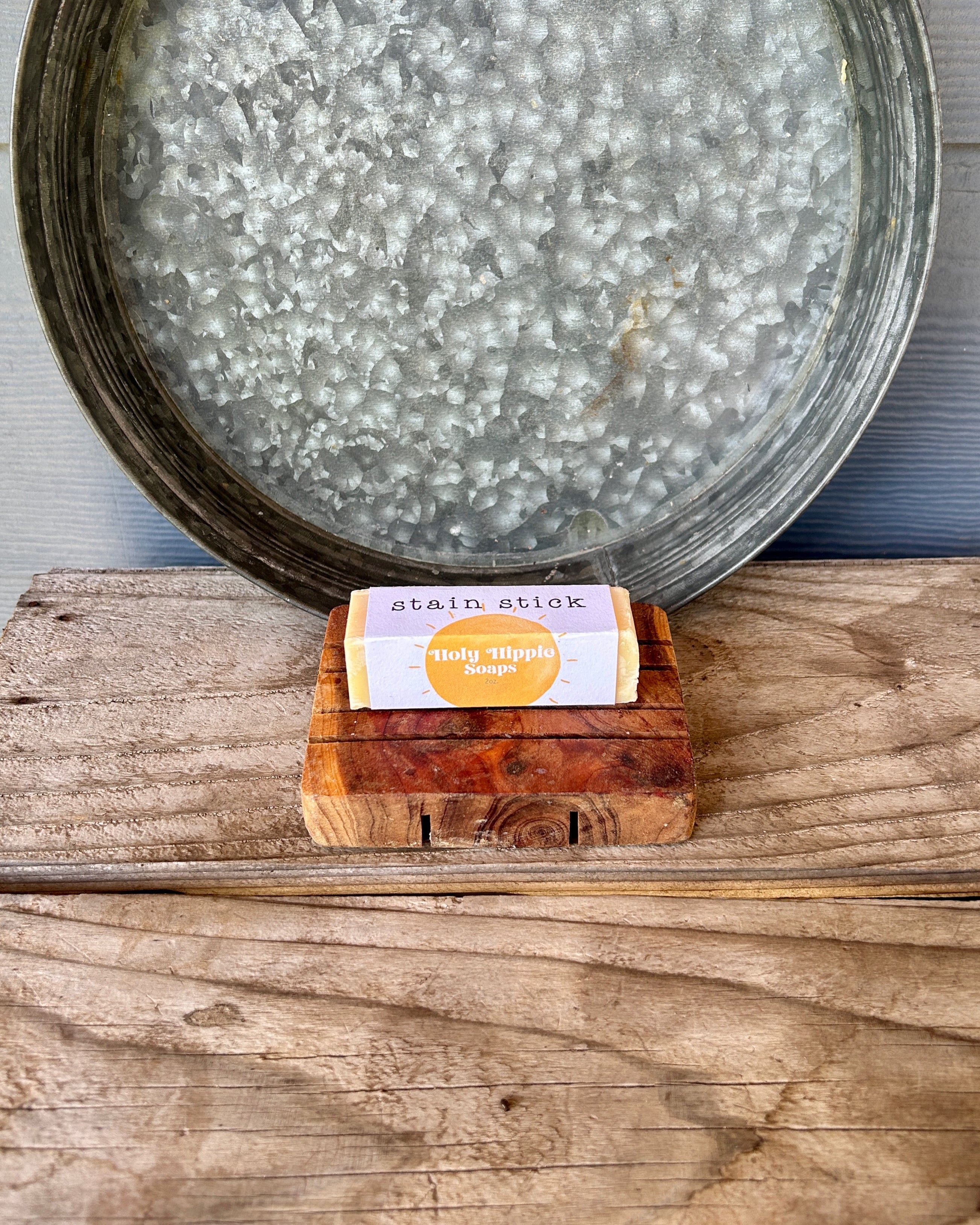 A packaged bar of stain-stick sits on a small wooden soap dish on a rustic wooden surface, with a large galvanized metal tray behind it.