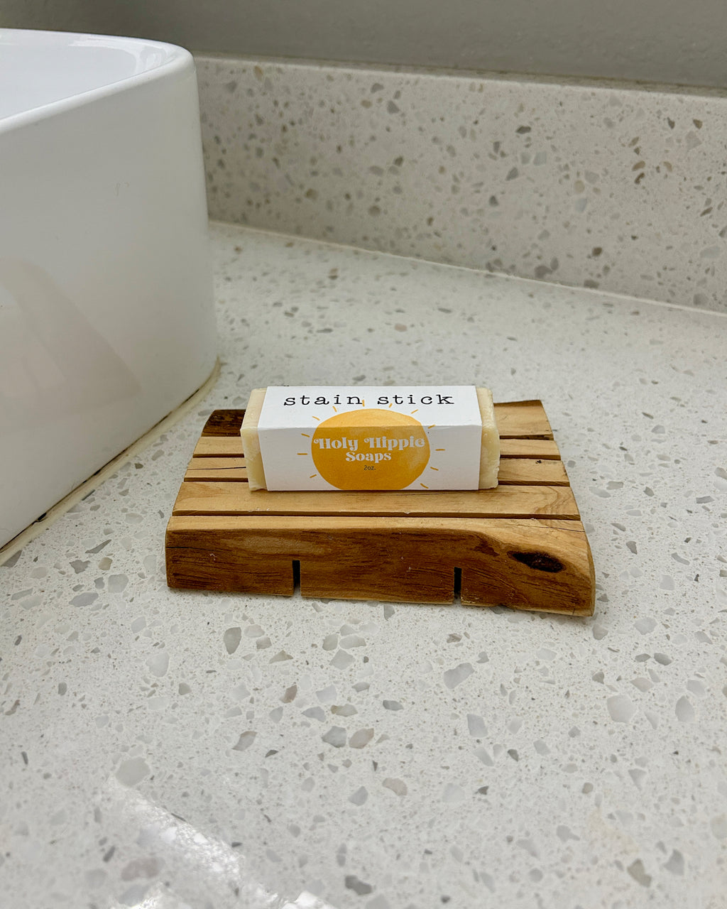 A packaged bar of Holy Hippie Soap 'stain stick' resting on a small wooden soap dish on a speckled bathroom countertop next to a white sink.