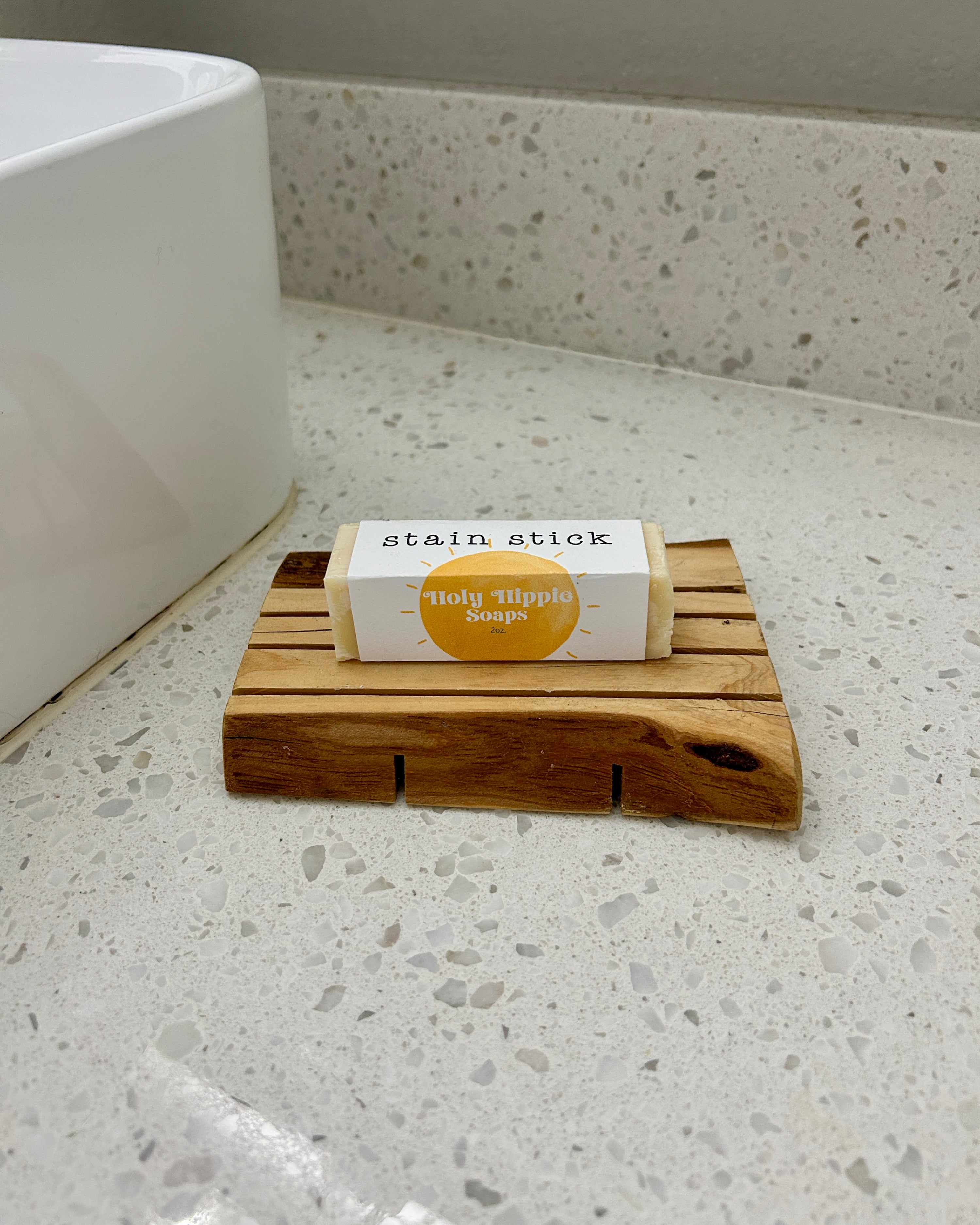 A packaged bar of Holy Hippie Soap 'stain stick' resting on a small wooden soap dish on a speckled bathroom countertop next to a white sink.
