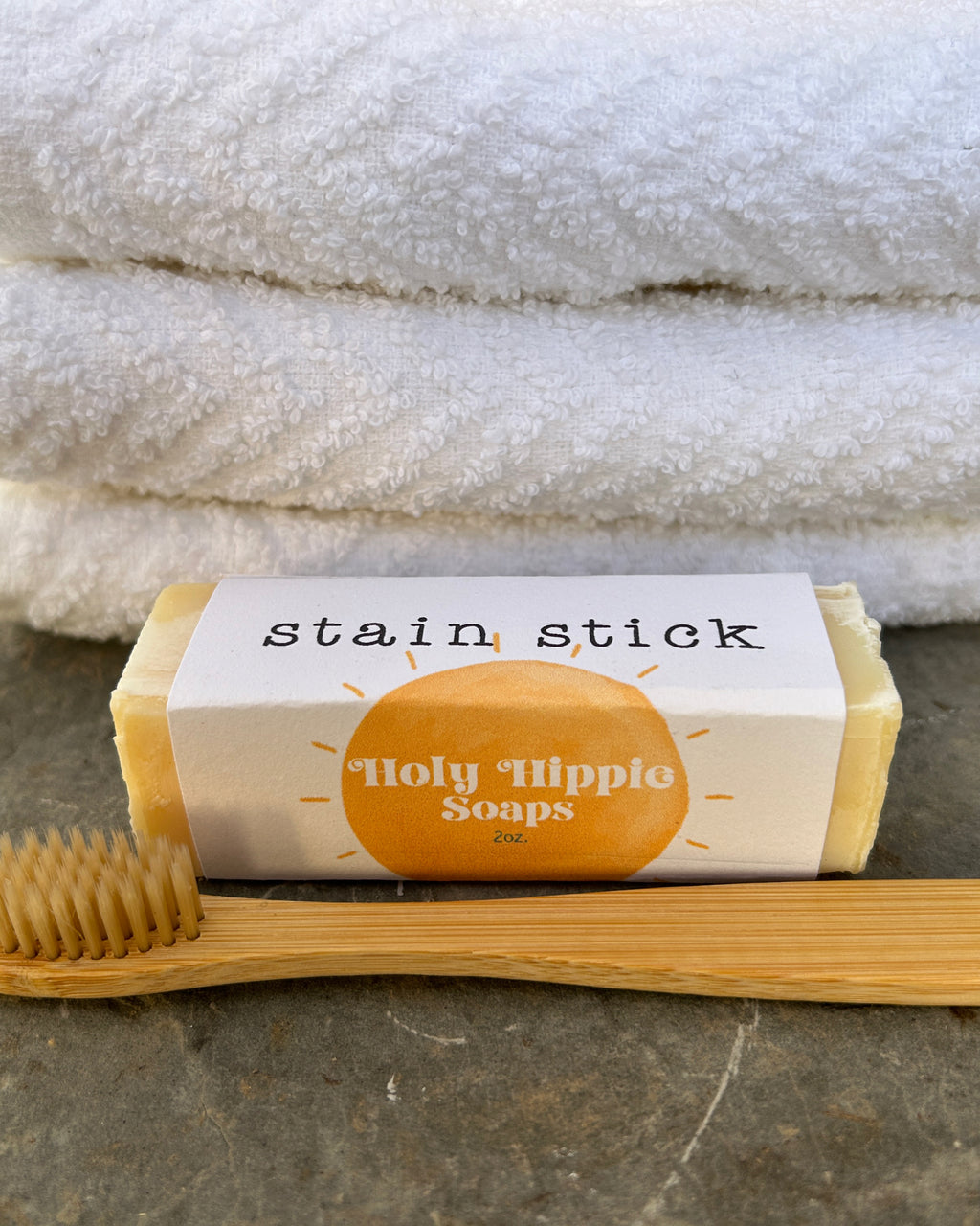A wrapped soap stain stick bar labeled 'Holy Hippie Soaps' sits on a stone surface with a bamboo brush in front and folded white towels behind it.